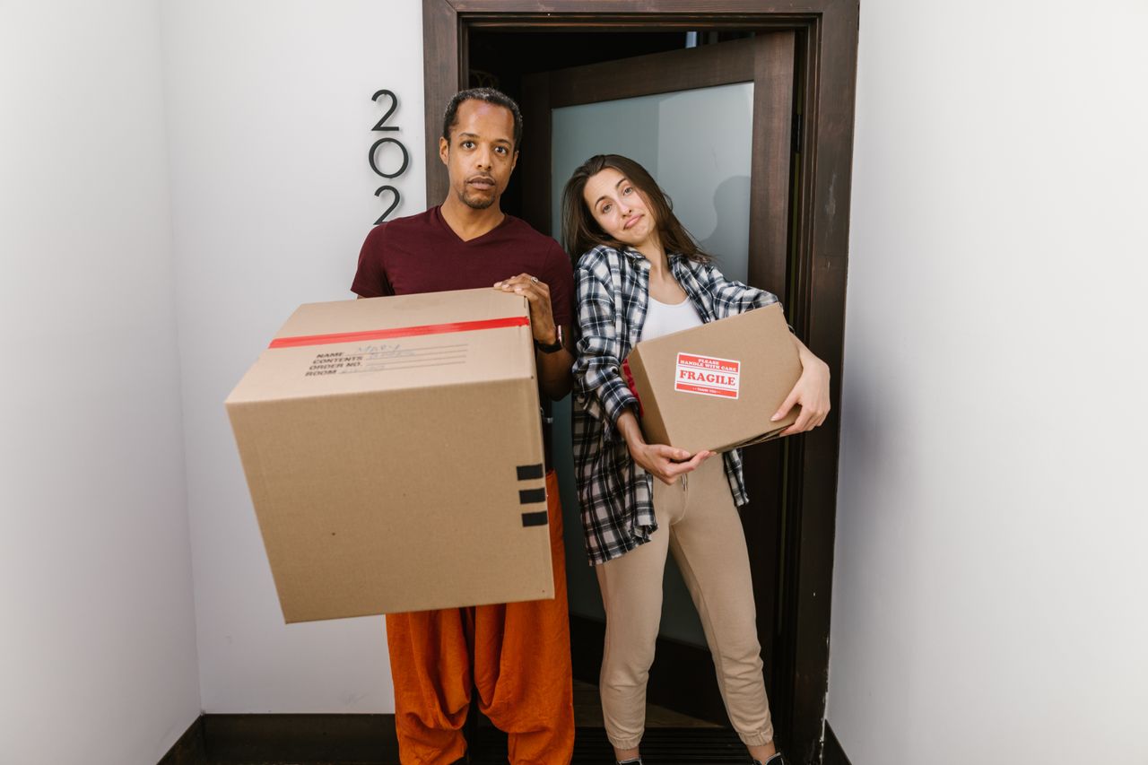 our-journey-01 A couple carrying moving boxes indoors, settling into their new apartment.